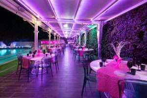 Terrazza ristorante all'aperto di sera con illuminazione viola e rosa, tavoli apparecchiati, parete verde e piscina sullo sfondo