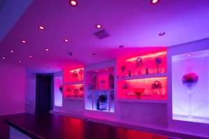 Interno di un locale con illuminazione LED rosa e viola, scaffali a muro con decorazioni e bancone in primo piano