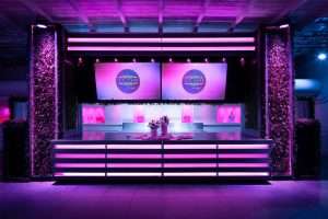 Stand espositivo J.T. Café con illuminazione viola e rosa, pareti verdi, due schermi con logo e bancone luminoso