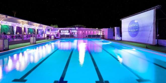 Piscina olimpionica del locale J.T. Cafe Eventi illuminata di notte al J.T. Café, con area lounge, bar e schermo di proiezione sullo sfondo